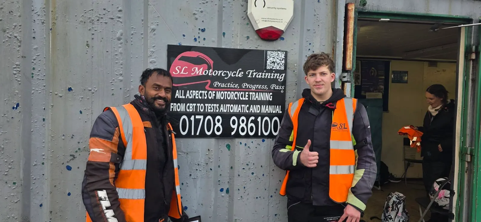 Two students in hi-vis vests giving a thumbs up in front of the SL Motorcycle Training sign