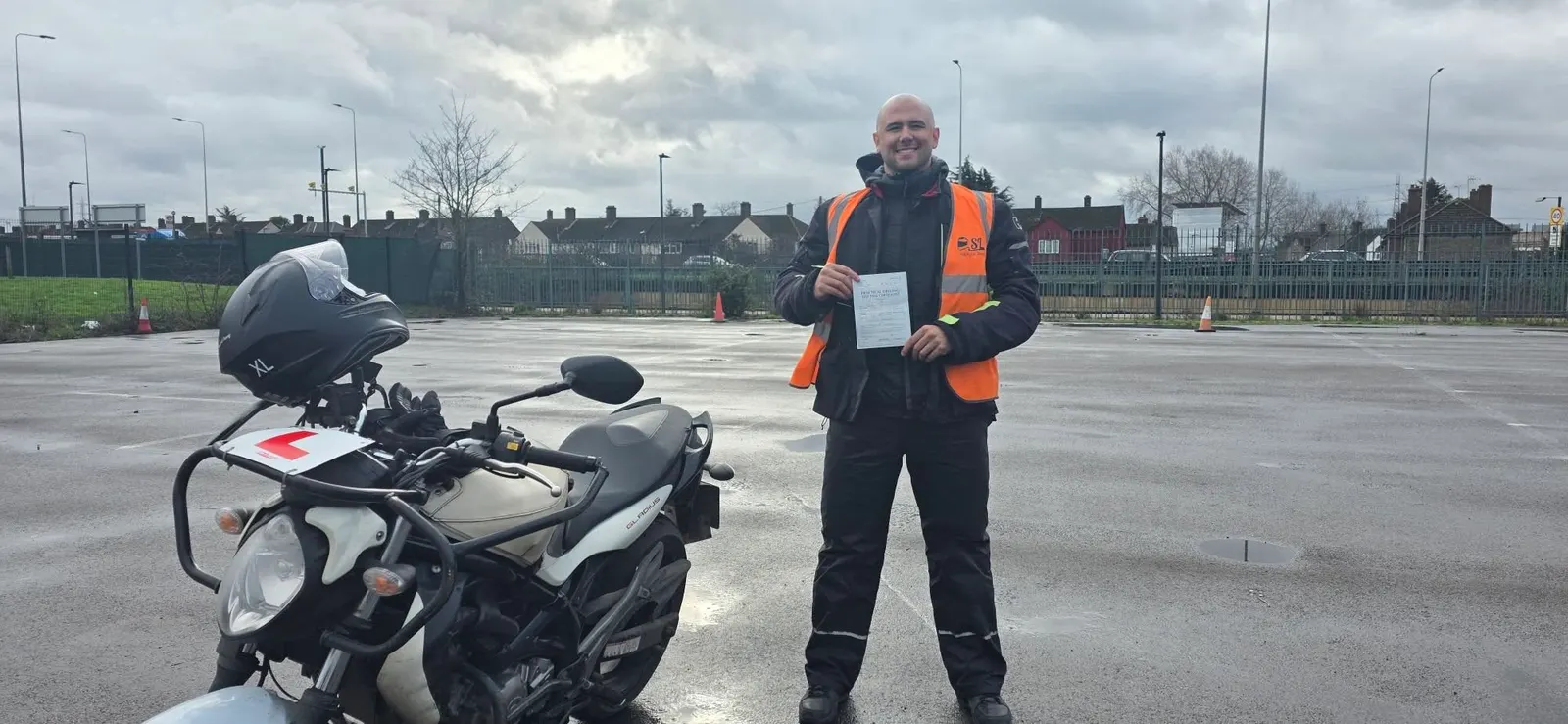 A student in an SL hi-vis vest holding his pass certificate on the training ground, next to a learner motorbike